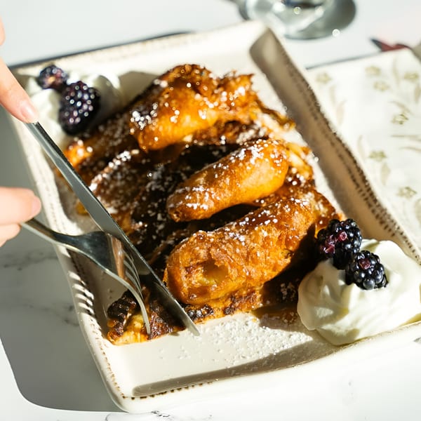 A person cuts into golden-brown French toast sticks dusted with powdered sugar, served with whipped cream and blackberries.