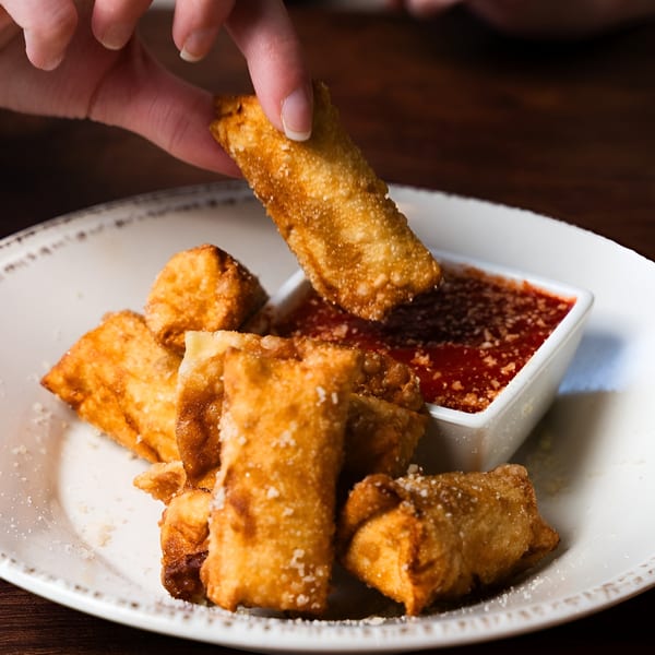 A hand dips a crispy, golden-brown egg roll into a small white bowl of red dipping sauce.