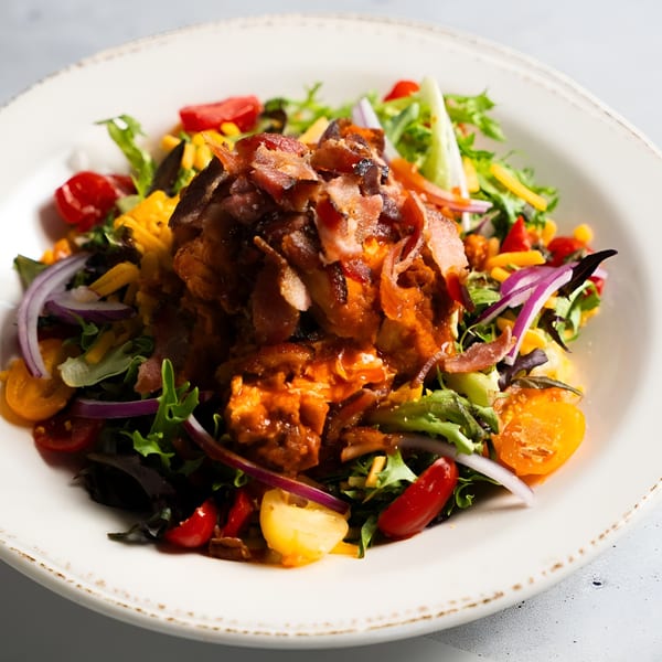 A BBQ chicken salad with mixed greens, tomatoes, red onion, cheese, and bacon in a white bowl.