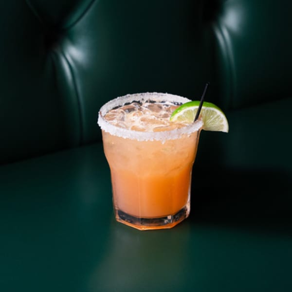 A salted margarita with a lime wedge and black straw, set against a dark green tufted bench.