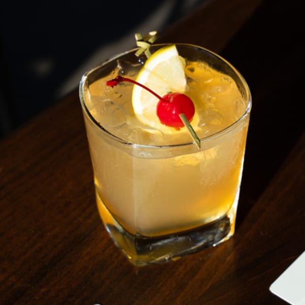 A whiskey sour cocktail with a lemon slice and a cherry on a dark wooden table.