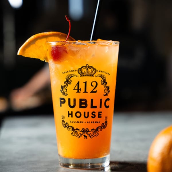 A vibrant orange cocktail with ice, garnished with an orange slice and cherry, featuring the 412 Public House logo.