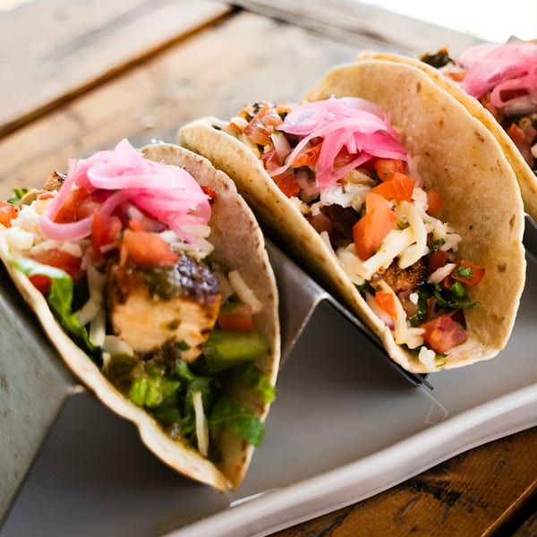 Three chicken tacos with pickled red onions, diced tomatoes, and shredded cheese on a wooden table.