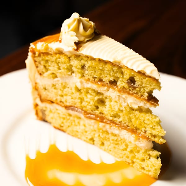 A slice of layered cake with white frosting and caramel drizzle on a white plate.