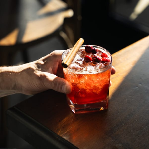 Hand holding a red cocktail with cranberries and a cinnamon stick on a dark wooden table.